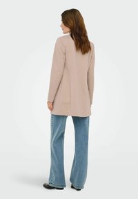 Veste beige à manches longues avec une coupe droite, associée à un jean évasé bleu clair. Texture de tissu lisse, design simple et détails minimaux.