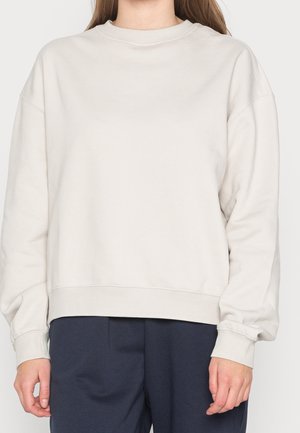 Person wearing a loose, long-sleeve beige sweatshirt paired with dark navy pants, standing against a plain background.