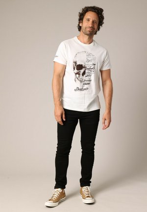 T-shirt en coton blanc avec un graphique de skull noir et des textes, associé à un jean noir et des baskets marron, se tenant devant un fond neutre.