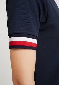 Granatowa koszulka polo z teksturowanej powierzchni. Rękaw z krótkim rękawem zdobiony jest czerwono-białymi pasami. Zbliżenie na część rękawa.