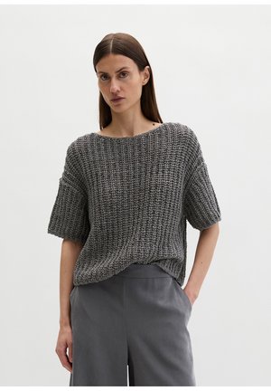 Frau mit glattem braunem Haar, die einen lockeren grauen, kurzärmeligen Strickpullover und hoch taillierte graue Hosen trägt, steht mit einer Hand in der Tasche.