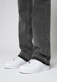 Piernas con jeans anchos negros desgastados con dobladillos deshilachados, combinados con zapatillas bajas blancas y limpias sobre un fondo liso.