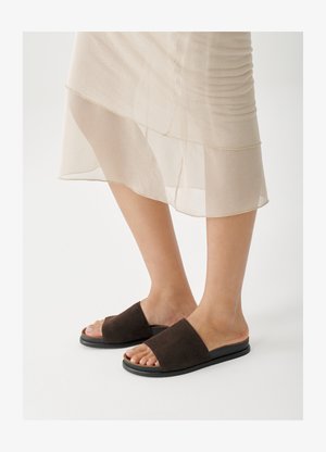 Sandale cu talpă joasă din pâslă maro, cu o curea lată, talpă neagră de cauciuc și design cu degetele descoperite, pe un fundal de culoare deschisă.