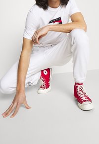 Röda högskurna sneakers med vita snören, prydda med en logotyp. Modellen bär en vit t-shirt och matchande träningsoverallsbyxor.