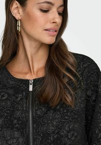 Črn jopič s cvetličnim vzorcem in teksturirano površino, ki ima srebrno zadrgo in okroglo ovratnik. Model ima dolge lase in velike uhane iz krogcev.