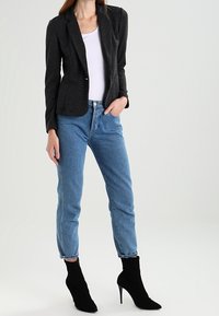Blazer noir ajusté, haut blanc, jeans taille haute bleus et bottines noires en chaussette. Tissu lisse, design ajusté et détails subtils sur le blazer.