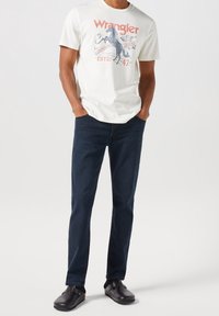 Man som bär en vit Wrangler grafik T-shirt, mörkblå jeans, svarta strumpor och svarta slip-on skor, står med händerna i fickorna.