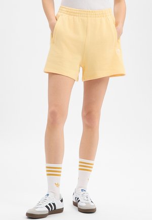 Legs wearing pale yellow shorts with hands in pockets, white Adidas socks with yellow stripes, and white Adidas sneakers with black stripes.