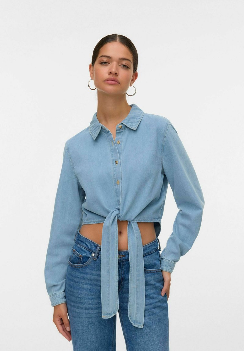 Camicia in denim azzurro chiaro, taglio cropped, con maniche lunghe, chiusura a bottoni frontale e dettaglio a nodo in vita. Presenta un colletto classico e polsini.