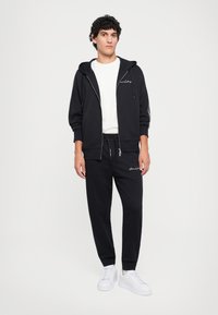 Young man wearing black zip-up hoodie, matching black joggers, white t-shirt, and white sneakers standing against a plain background.