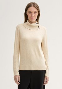 Beżowy sweter z golfem o prążkowanym kołnierzu, z dwoma czarnymi guzikami po lewej stronie, wykonany z miękkiej, teksturowanej tkaniny.
