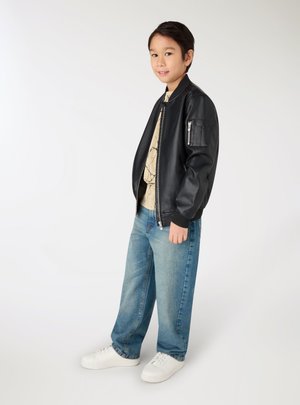 Ragazzo giovane con giacca di pelle nera, camicia chiara a fantasia, jeans blu e scarpe da ginnastica bianche, in piedi su sfondo bianco.