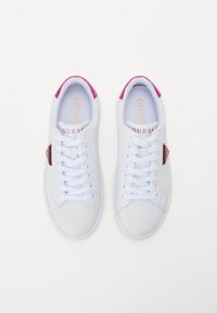 Valkoiset lenkkitossut, joissa on sileä synteettinen päällinen, vaaleanpunaiset yksityiskohdat kantapäässä, pyöreä kärki ja perinteinen nauhoitus. Kielen päällä kultainen logo.