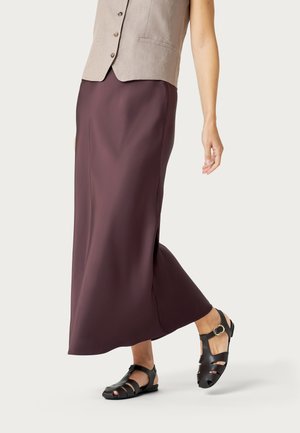 Mujer vestida con un chaleco beige, falda larga de color púrpura oscuro y sandalias negras abiertas con tiras en el tobillo, de pie contra un fondo liso.