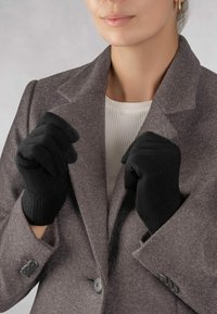 Schwarze Strickhandschuhe mit gerippten Bündchen, getragen von einer Person in einem grauen Wollmantel, die die Hände vor dem Oberkörper geballt hat.