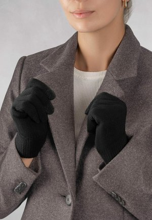 Gants en tricot noirs avec des poignets côtelés, portés par une personne dans un manteau en laine gris, les mains serrées devant le torse.