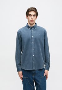 Modrá kordurová košeľa na gombíky s golierom, dlhými rukávmi a malým logom na hrudi. Vrejdová textúra a biele gombíky.