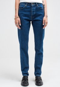 Jeans de mezclilla azul con corte recto y talle medio. Presenta un diseño clásico de cinco bolsillos y cremallera. Costuras detalladas visibles.