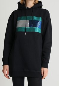 Czarna bluza z kapturem z frontalnym wzorem z cekinów, zawierającym niebieskie, zielone, srebrne i szare paski. Regulowany sznurek i ściągacze przy mankietach.