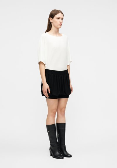 Γυναίκα με μακριά καστανά μαλλιά, φοράει άσπρο φαρδύ τοπ, μαύρη μίνι φούστα με πιέτες και μαύρες δερμάτινες μπότες μέχρι το γόνατο, στέκεται μπροστά σε γκρι φόντο.