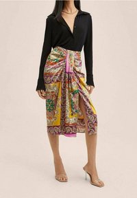 Chemise noire à manches longues associée à une jupe à motifs colorés présentant des teintes jaunes, roses et vertes, avec un design drapé et une fente sur le côté.