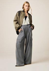 Olivengroene bomberjack met zwarte borduring, witte overhemd met knopen, en losse grijze cargo pants met studs in de zakken en een elastische taille.
