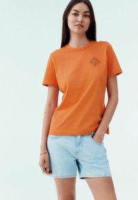 Oranssi puuvillapaita, jossa on lyhyet hihat ja pyöreä kaula-aukko, jossa on pieni musta logo rinnassa, yhdistettynä vaaleansinisiin farkkushortseihin.