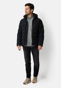 Schwarze Steppjacke mit Kapuze, strukturierter Optik, mit einer Brusttasche, getragen über einem hellgrauen Hemd mit Knöpfen und schwarzen Jeans.
