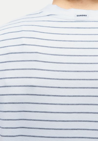 Κοντινή λήψη λευκού υφάσματος με λεπτές οριζόντιες ναυτικές ρίγες και μια μικρή ετικέτα "Sandro" στο πίσω μέρος του λαιμού στην κορυφή.