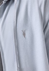 Világoskék, cipzáros kapucnis pulóver puha anyagból. A bal mellrészen egy kis, szürke hímzett kecskebőr koponya logó található.