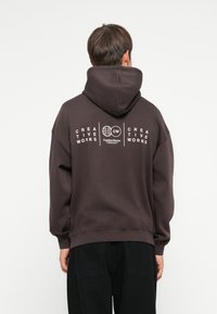 Brun hætte sweatshirt med en afslappet pasform, der har hvid trykt tekst og et globusdesign på ryggen. Blød stofstruktur.