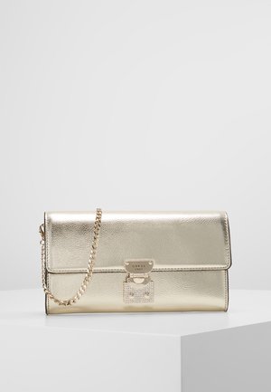 Guldmetallic clutch-taske med kæderem, frontlås indgraveret med "GUESS 1981," hvilende på en hvid piedestal.