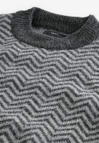 Grauer Strickpullover mit einem Zickzackmuster in verschiedenen Grautönen. Die strukturierte, dicke Kragen sorgt für Detail und Wärme. Markenschild sichtbar.