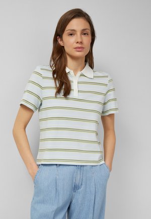 Kobieta o brązowych włosach ubrana w biało - jasnozieloną koszulkę polo w paski i jasnoniebieskie, jeansy z wysokim stanem, z rękami w kieszeniach.