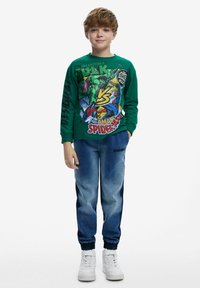 Grön långärmad skjorta med grafik av "Incredible Hulk" och "Spider-Man". Matchad med blå denim joggers och vita sneakers.