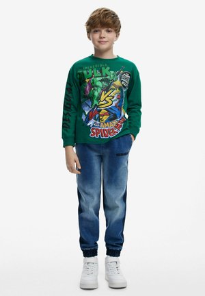 Zelena košulja dugih rukava s grafikom "Nevjerojatni Hulk" i "Spider-Man". Uparena s plavim trapericama i bijelim tenisicama.