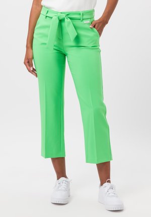 Green tailored pants with a tie belt, cropped length, smooth texture, fitted waist, and side pockets, paired with white sneakers.