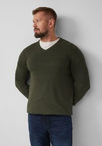 Olivgrüner, langärmeliger V-Ausschnitt-Pullover mit strukturiertem Strick, mit einem dezenten Logo auf der Brust, getragen über einem weißen T-Shirt.