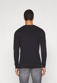 Maglione nero a maniche lunghe realizzato in tessuto a maglia testurizzata. Presenta un colletto rotondo e polsini e bordo a coste. Indossato con jeans grigi.