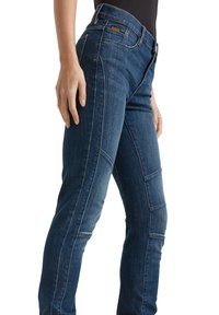 Kvinna som bär mörkblå denimjeans med synliga sömdetaljer och en liten brun logopatch nära framfickan.