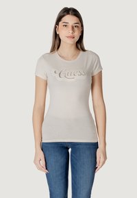 Camiseta ajustada de color beige claro con mangas cortas, que presenta un logo brillante de "Guess" en el pecho. Combinada con jeans azules.