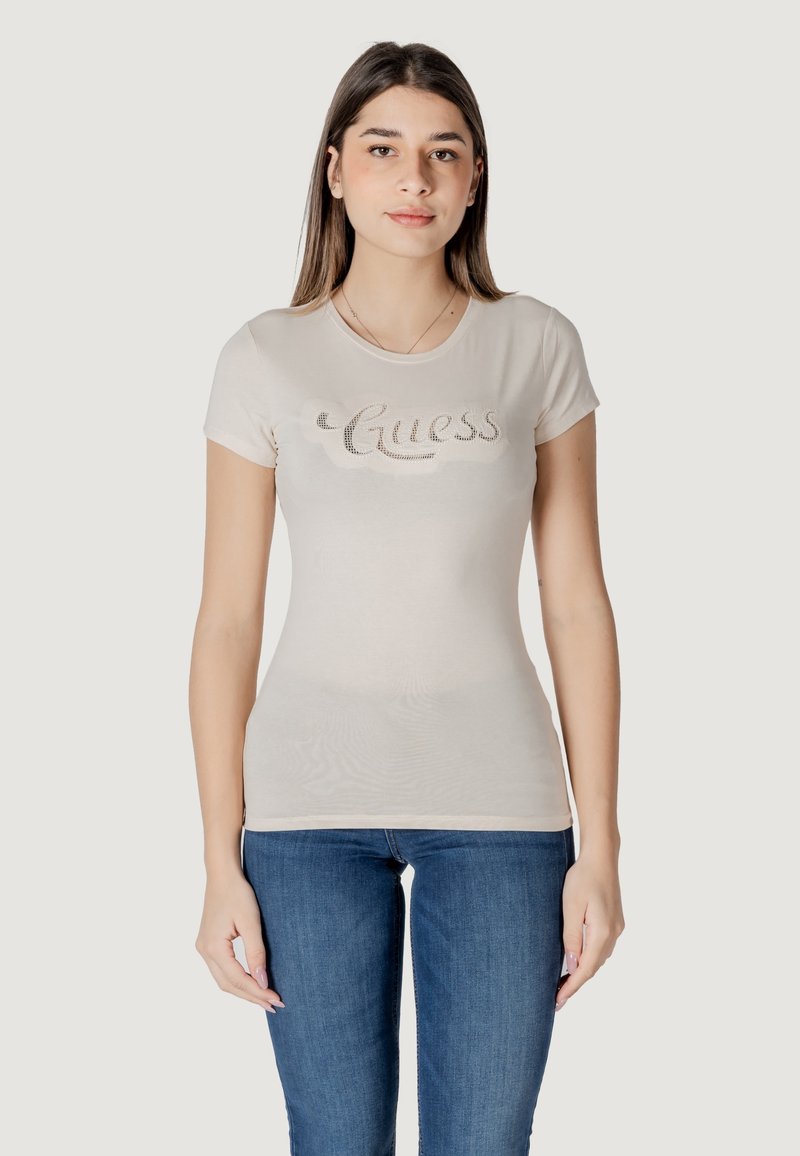 Camiseta ajustada de color beige claro con mangas cortas, que presenta un logo brillante de "Guess" en el pecho. Combinada con jeans azules.