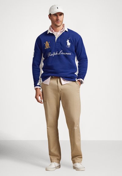 Άνδρας που φοράει άσπρο καπέλο, μπλε ράγκμπι πουκάμισο Ralph Lauren πάνω από ροζ πουκάμισο με γιακά, μπεζ παντελόνι και άσπρα αθλητικά παπούτσια, στέκεται μπροστά σε απλό φόντο.