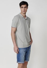 Svetlo siva polo majica s kratkimi rokavi, z dvema gumboma na zapenjanju in diskretnim logotipom na prsih. V kombinaciji z jeans kratkimi hlačami z zavihanimi robovi.
