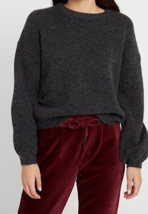 Femme portant un pull gris foncé en tissu duveteux à manches longues et un pantalon en velours côtelé bordeaux taille haute, noué avec un nœud à la taille.