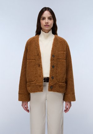 Vrouw met een bruine, pluizige jas over een witte coltrui en lichte broek, staand tegen een effen lichte achtergrond.
