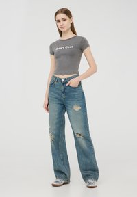 T-shirt grigio corto con testo "Non mi interessa", abbinato a jeans larghi blu strappati e scarpe argentate. Design semplice con strappi visibili.