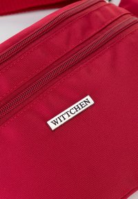 Bolsa de tela roja con múltiples compartimentos con cremallera. Presenta una placa de logo rectangular plateada que dice "WITTCHEN". Se puede ver un detalle de superficie texturizada.