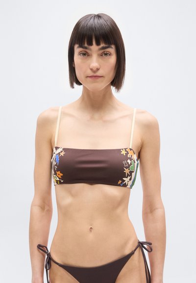 Femme aux cheveux courts et foncés portant un bikini marron avec des imprimés floraux et animaliers sur le haut et un bas noué, debout devant un fond uni.