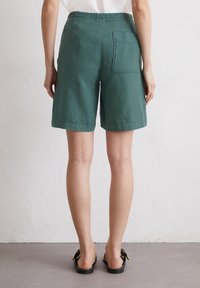 Shorts en lin verts, longueur genou, avec une poche arrière. Texture lisse, taille large et ourlet replié. Portés avec des sandales plates noires.